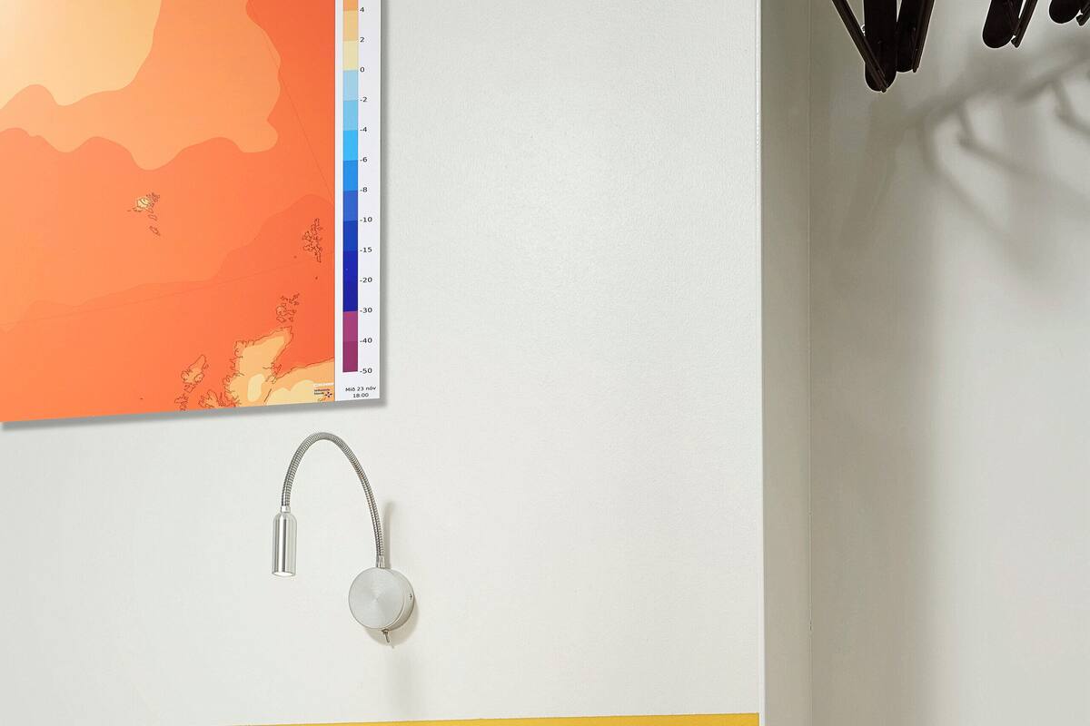 A hotel room with a bed, a small table with a telephone and a remote control, a wall-mounted lamp, and a colorful map on the wall.