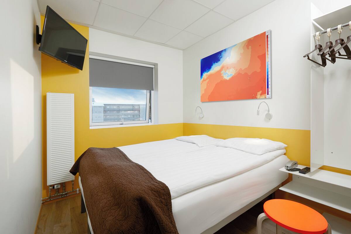 A hotel room with a bed, a window, a flat-screen TV, a painting, a towel rack, and a sink.