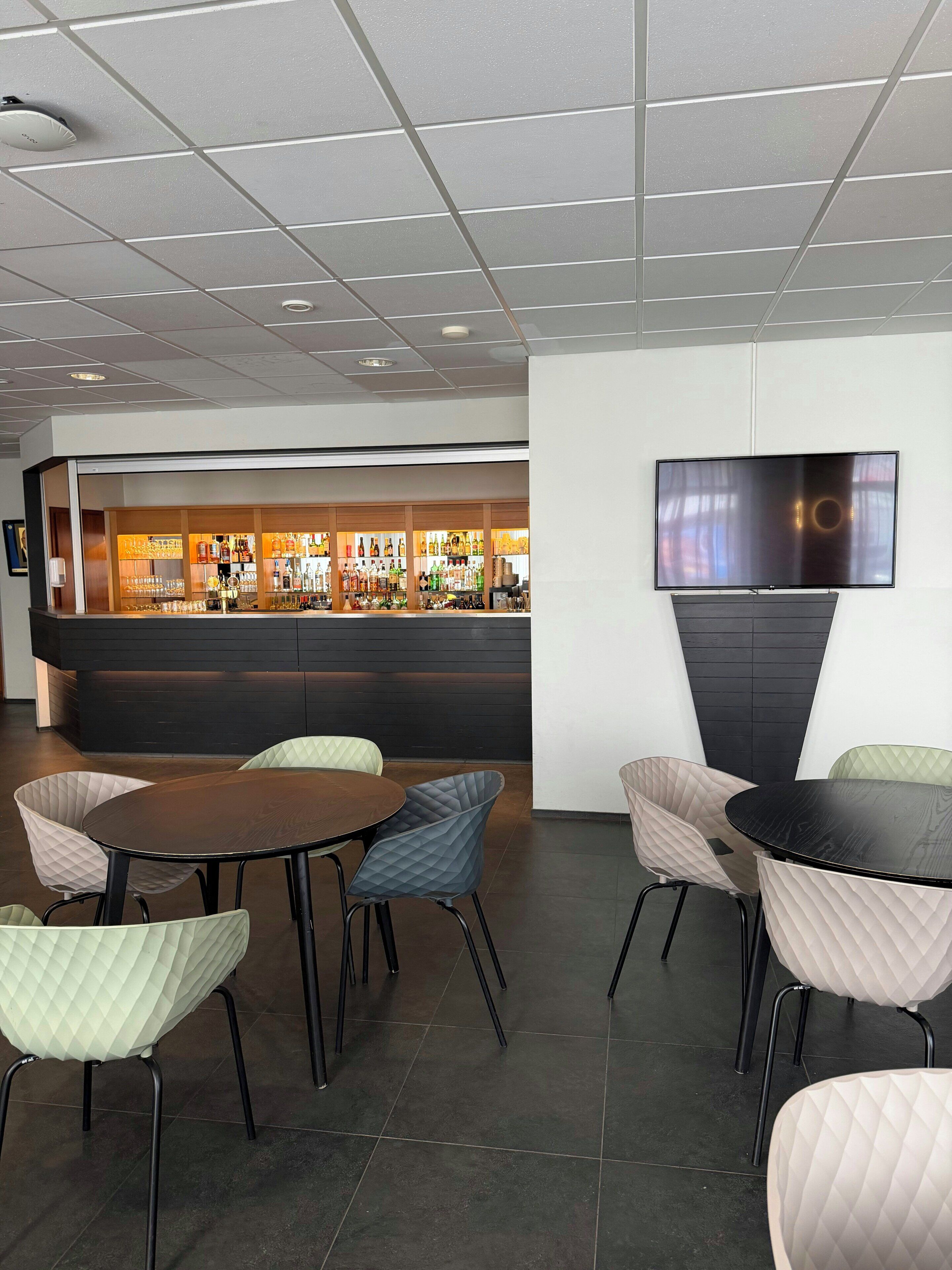 A bar area with a flat-screen TV mounted on the wall, a bar counter with various bottles and glasses, and several cushioned chairs around a round table.