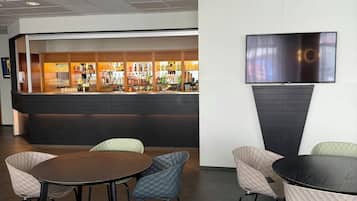 A bar area with a flat-screen TV mounted on the wall, a bar counter with various bottles and glasses, and several cushioned chairs around a round table.