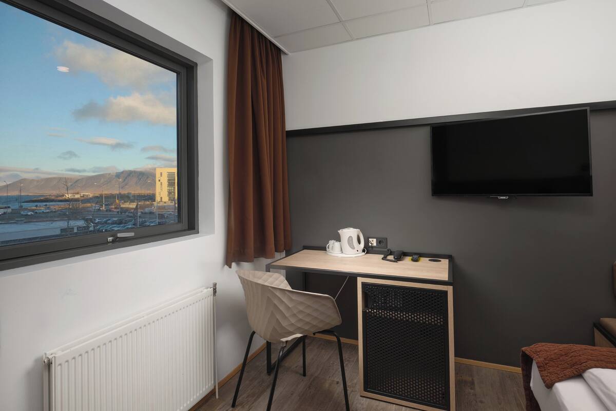 Quarto de hotel com uma janela grande, uma escrivaninha com chaleira, uma cadeira, uma cama e uma televisão fixada na parede.