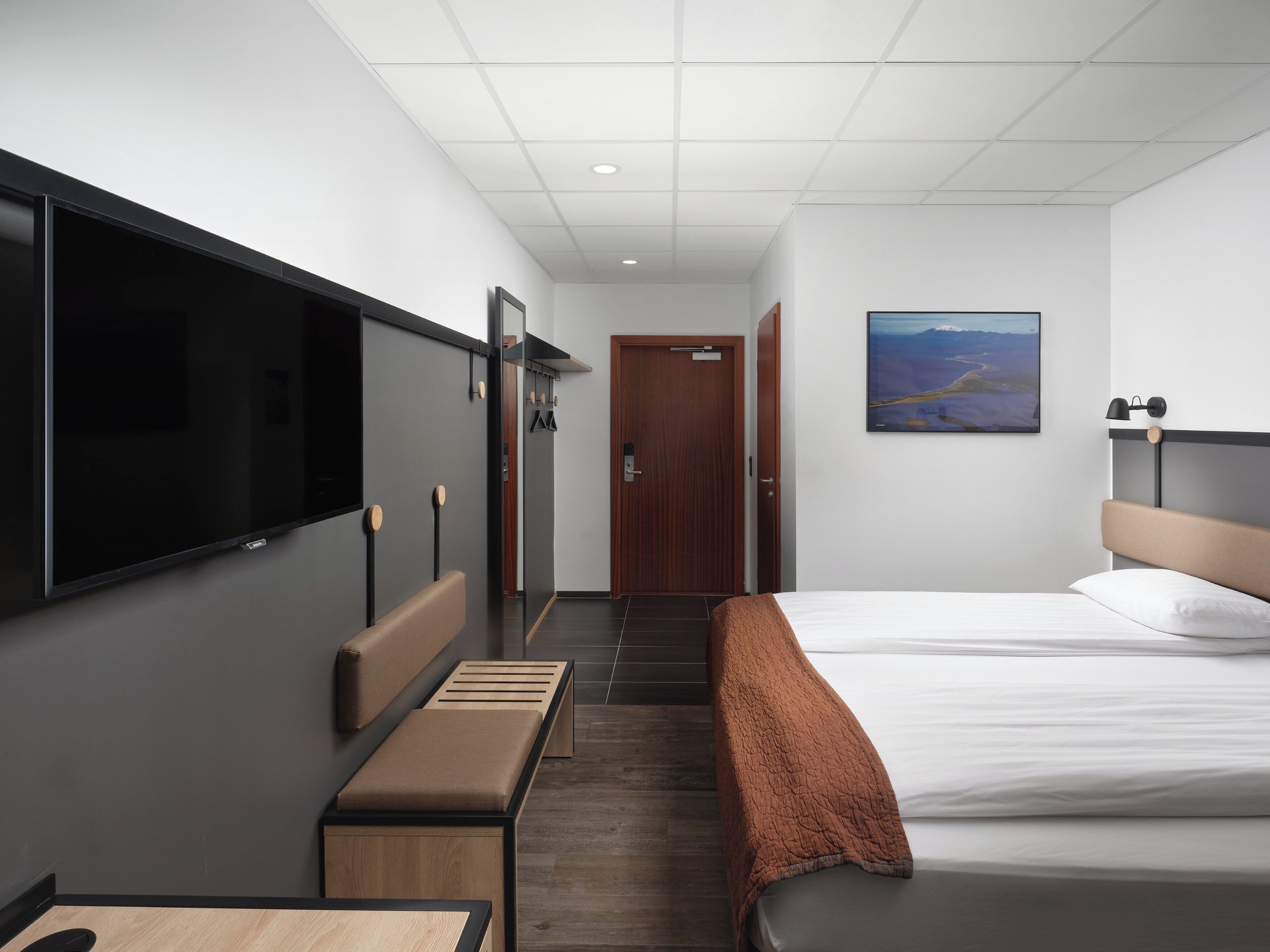 A hotel room with a bed, a TV, a bench, a door, and a painting on the wall.
