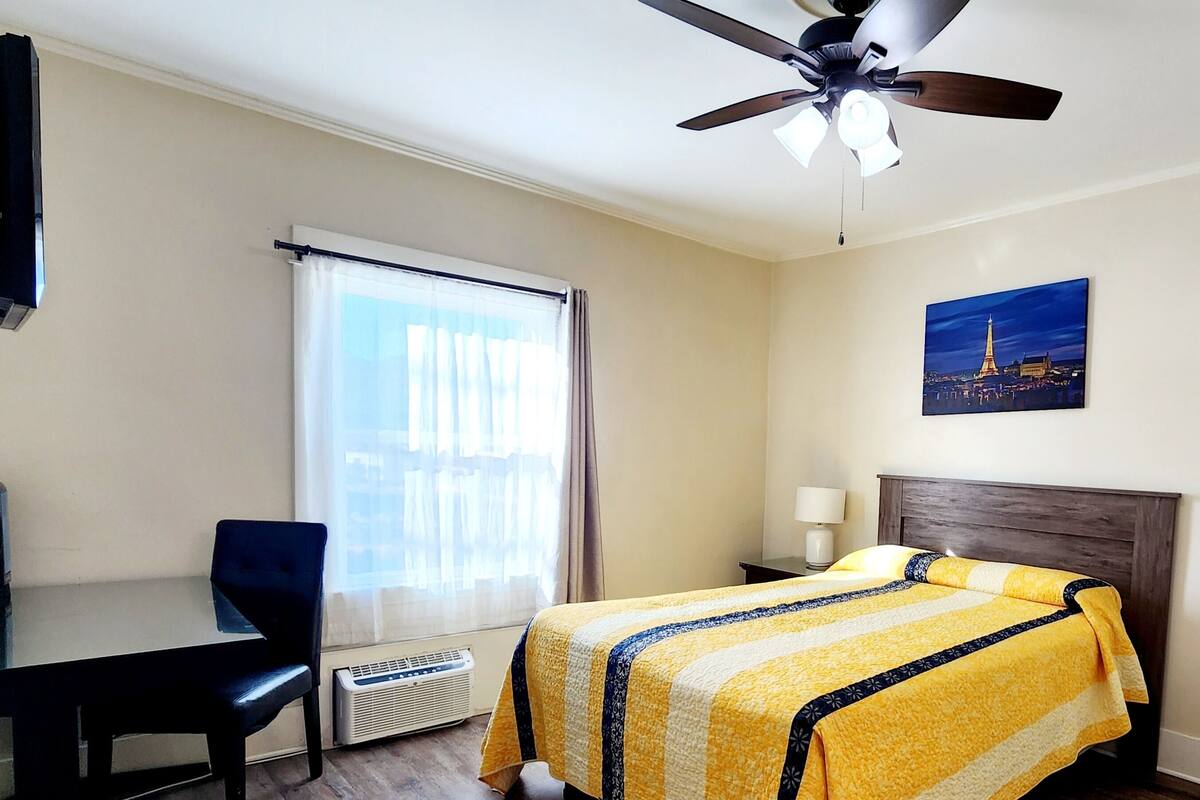 A hotel room with a bed, a desk, a chair, a ceiling fan, a window with curtains, and a painting of a tower.