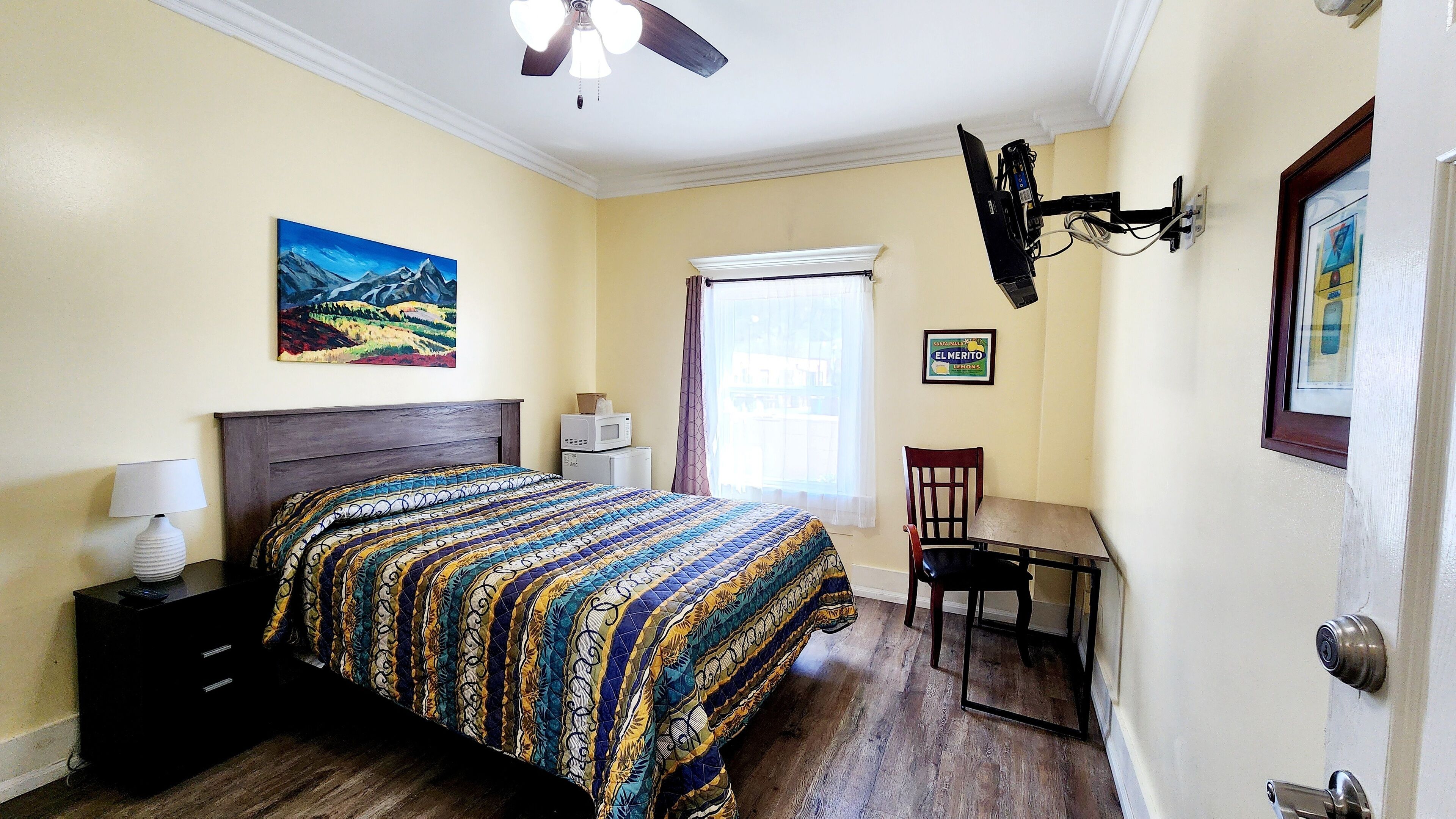 A bedroom with a bed, a desk, a chair, a ceiling fan, a window with curtains, and a painting on the wall.