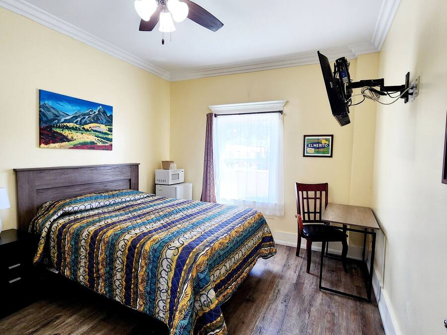 A bedroom with a bed, a desk, a chair, a ceiling fan, a window with curtains, and a painting on the wall.