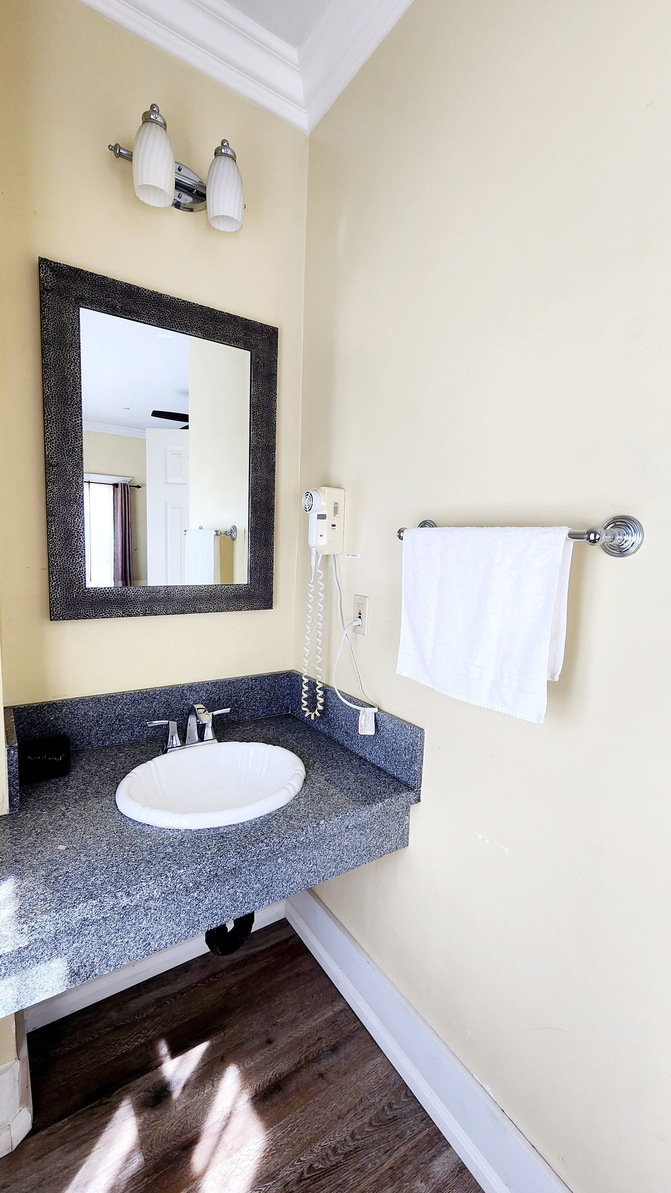 Un baño con lavamanos de encimera de granito, un espejo grande con marco negro, un perchero con una toalla blanca y un secador de pelo en la pared.