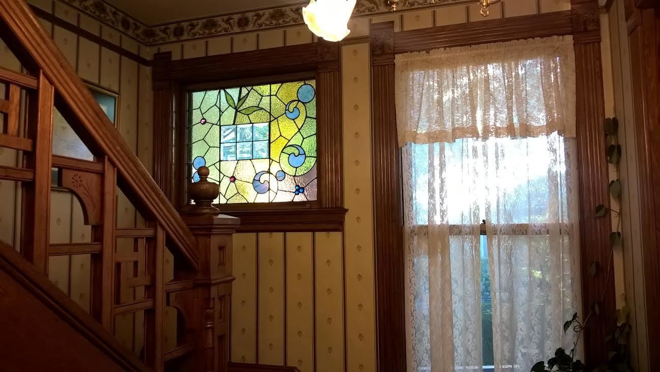 Una habitación con una escalera de madera, un vitral y un ventanal con cortinas de encaje.