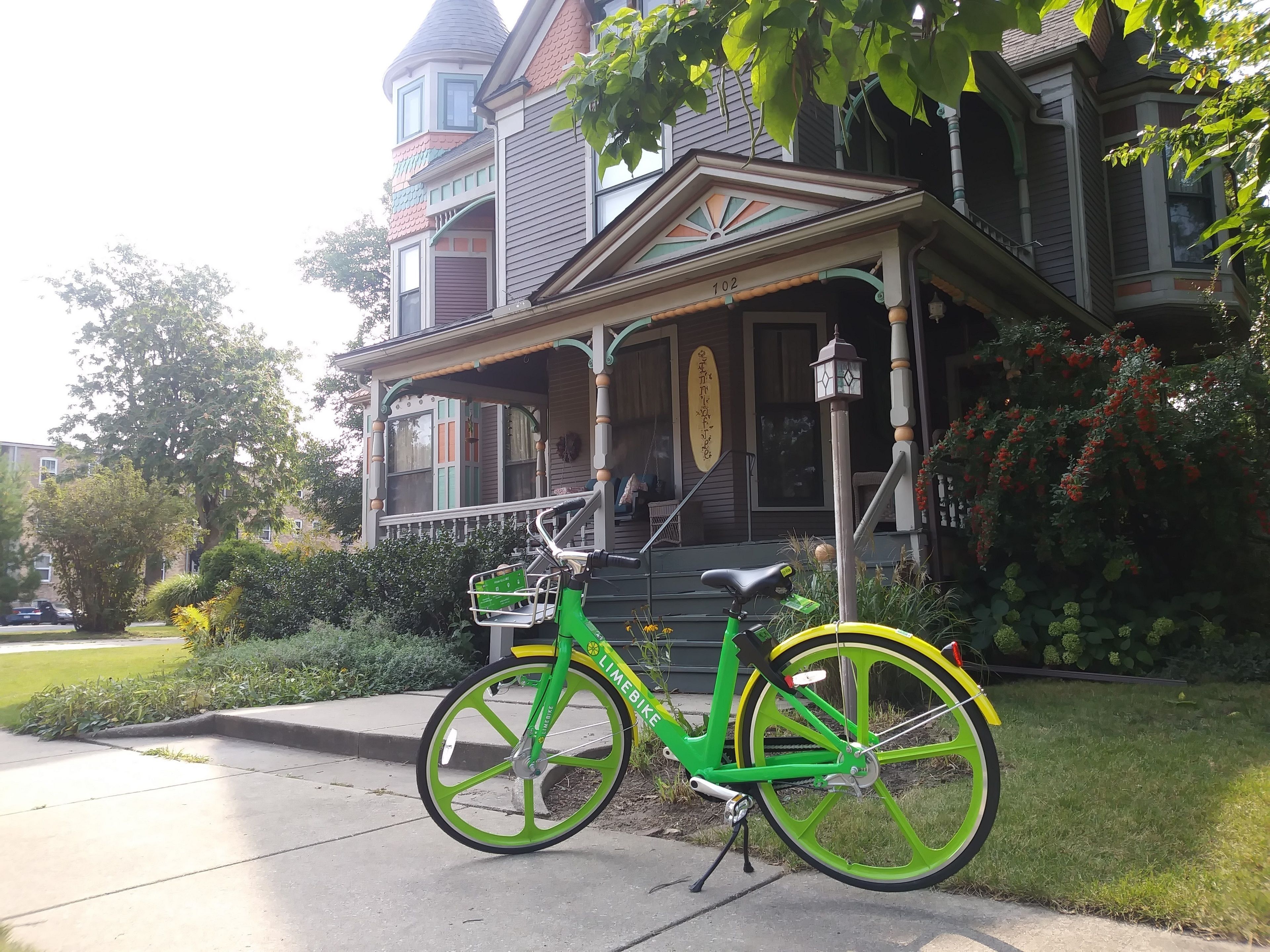 A green bicycle parked in front of a Victorian-style house with a porch and a number '102' sign.