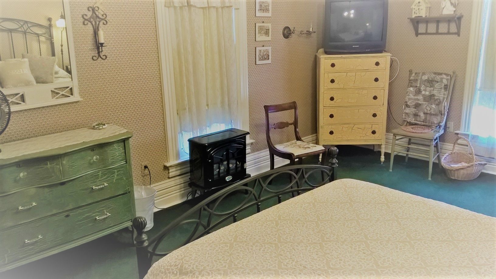 A room with a bed, a green dresser, a wooden chair, a small table with a radio, and a television on a cabinet.