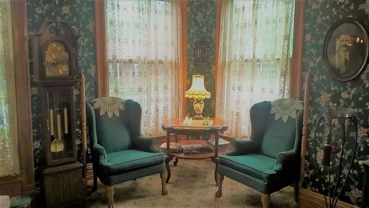 A room with two green armchairs facing a table with a lamp, a clock on the left, and floral wallpaper.