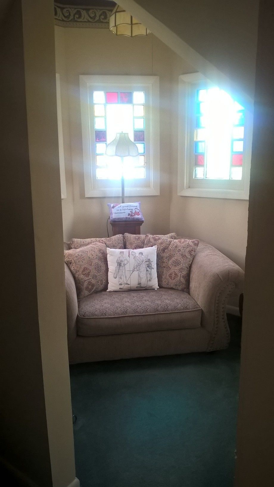 A room with a beige sofa, a small table with a lamp, and two windows with colorful stained glass.