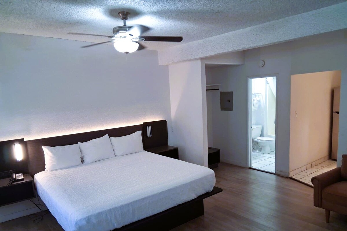 A bedroom with a large bed, a ceiling fan, a nightstand, and a door leading to a bathroom.