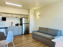 A compact kitchen with a stainless steel refrigerator, a microwave, and a coffee maker. There's a grey sofa and a white chair in the living area.