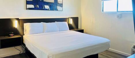Habitación de hotel con cama, mesitas de noche, ventilador de techo, una obra de arte colgada en la pared y una ventana con persianas.