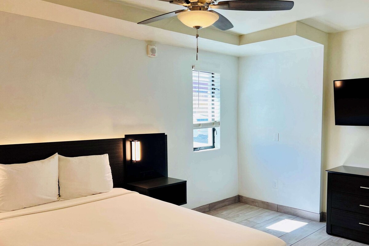 Une chambre d’hôtel comprenant un lit, une table de chevet, un ventilateur de plafond, une fenêtre avec des stores, un téléviseur et une armoire.