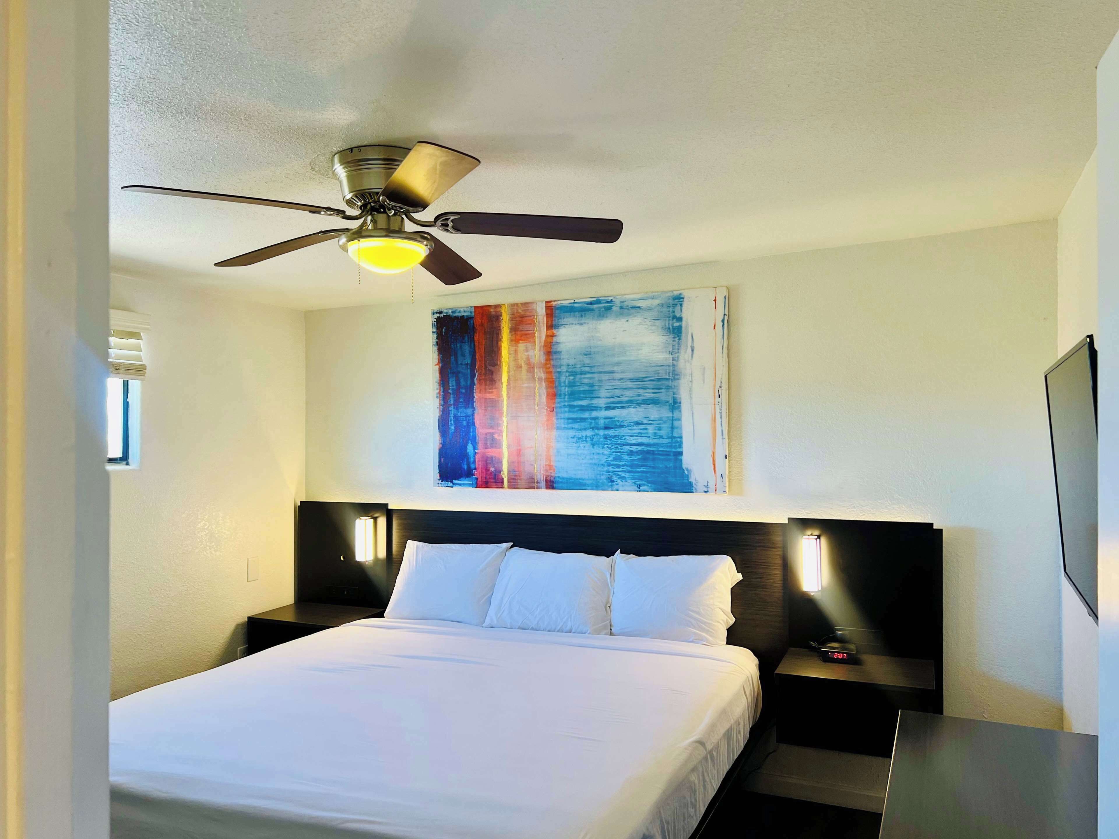 Una habitación de hotel con una cama, un ventilador de techo, un cuadro abstracto colgado en la pared y una mesita de noche con lámpara.