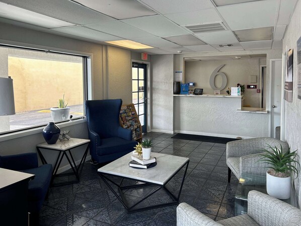 A reception area with a blue armchair, a small table with a book and a plant, and a reception desk with a computer and informational brochures. - Studio 6 Sierra Vista, AZ – Fort Huachuca (Sierra Vista)