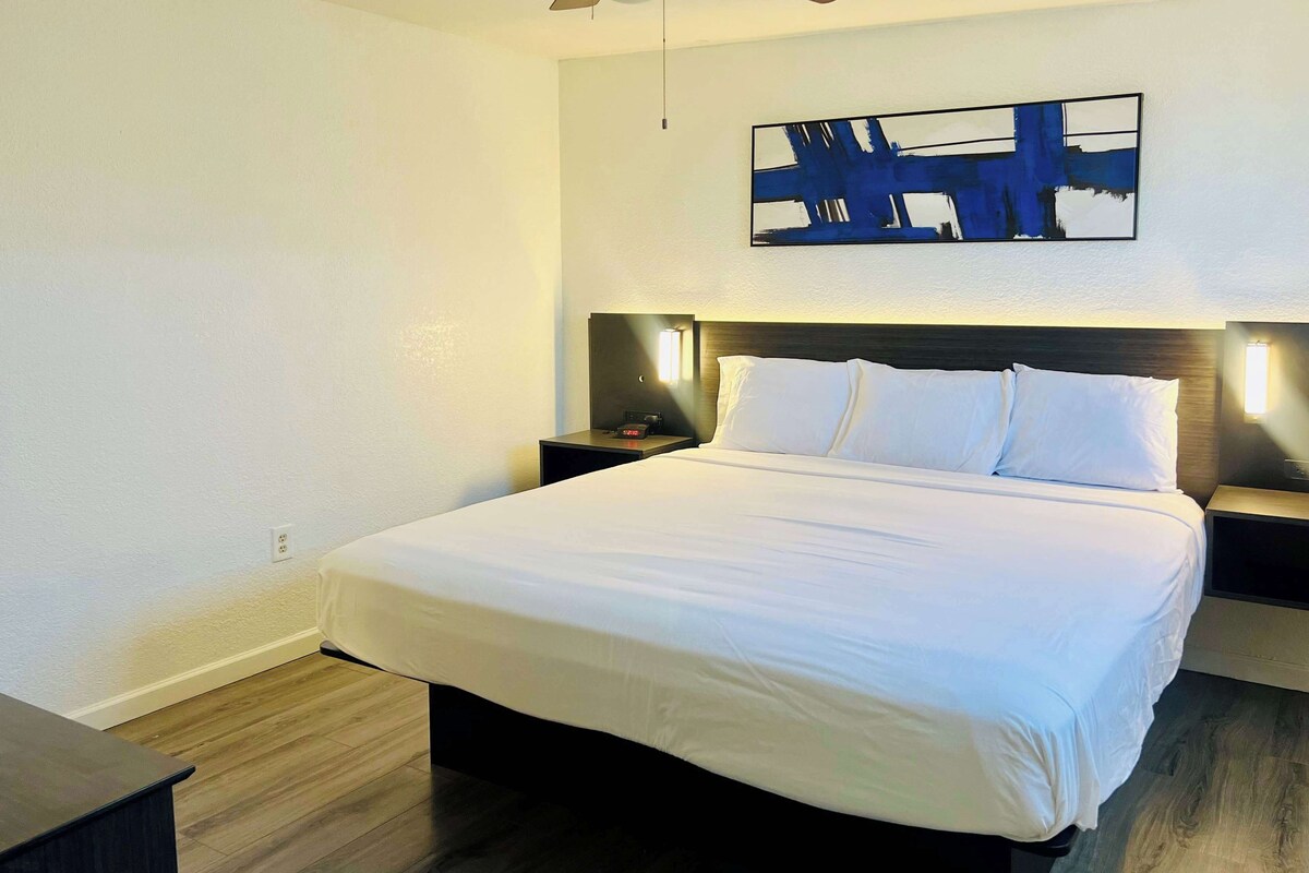 A neatly made bed with white linens, a dark wooden headboard, and a modern abstract painting above it.