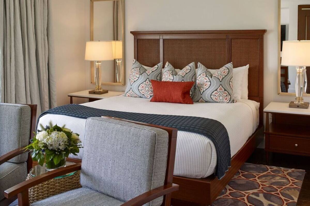 A hotel room with a large bed, two bedside tables with lamps, a wooden headboard, patterned pillows, a small table with a vase of flowers, and a chair.