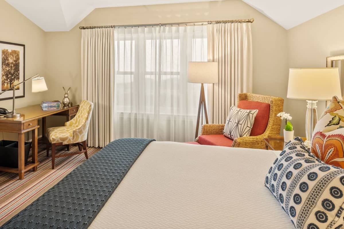 A hotel room with a large bed, a desk with a chair, a wardrobe, and a window with curtains.