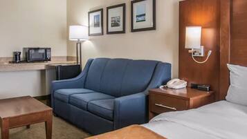 Habitación de hotel con una cama, un sofá azul, una mesita, una mesita de noche con lámpara, un microondas y cuadros en la pared.