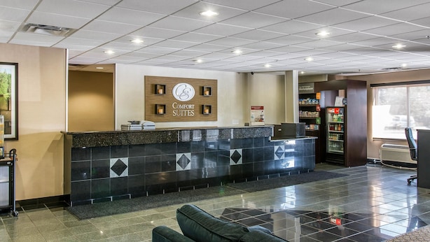 Uma área de recepção de hotel com balcão, placa com 'Comfort Suites', máquina de venda automática e espaço de estar com sofá.