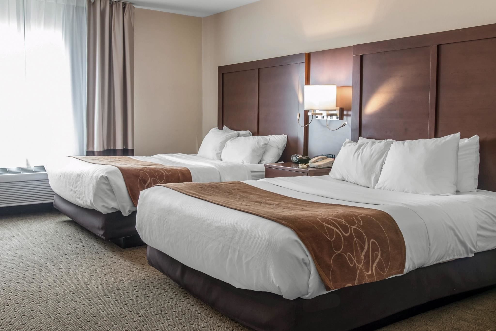 A hotel room with two beds, a large mirror, a bedside table with a lamp, and an air conditioning unit.