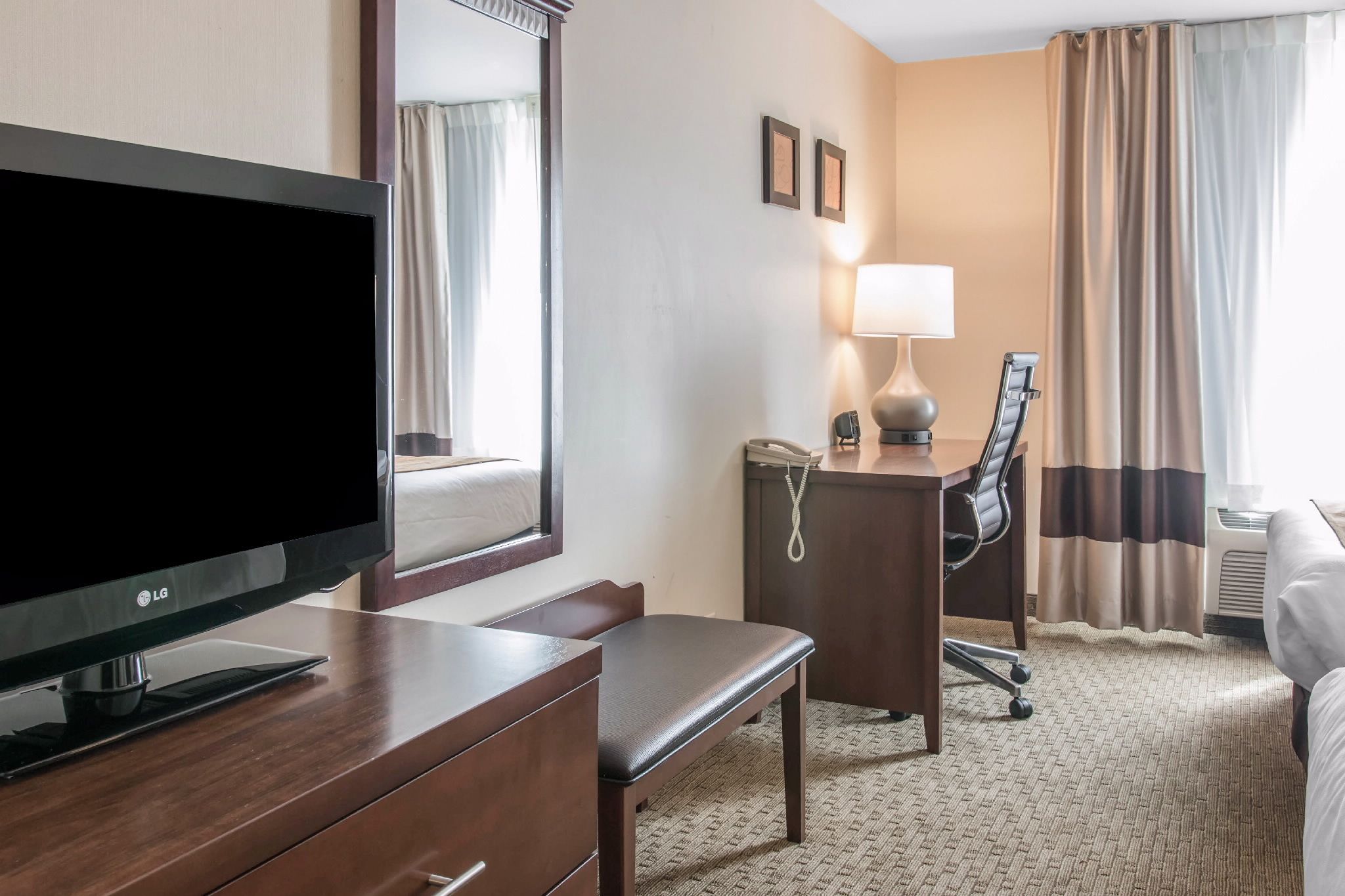 Habitación de hotel con televisión de pantalla plana, escritorio con lámpara, silla, cama y ventana grande con cortinas.