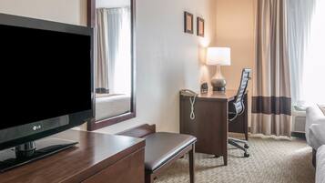 Une chambre d’hôtel équipée d’une télévision à écran plat, d’un bureau avec une lampe, d’une chaise, d’un lit et d’une grande fenêtre avec des rideaux.