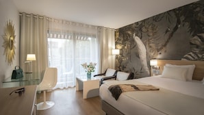 A hotel room with a large bed, a desk with a chair, a glass table, a lamp, and a wall mural. - Hotel Chambord (Menton)