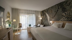 A hotel room with a large bed, a desk with a chair, a small table with a lamp, and a large window with curtains. - Hotel Chambord (Menton)