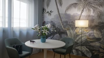 Una habitación con una mesa redonda, dos sillas, un florero con flores, una lámpara y un mural con follaje tropical en la pared.
