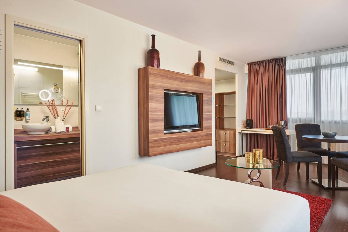 A modern hotel room with a large bed, a TV mounted on a wooden panel, a desk with a chair, and a dining area with a table and chairs.