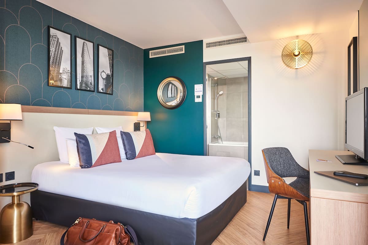A hotel room with a large bed, a desk with a chair, a television, and a bathroom visible through an open door.