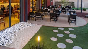 Un patio al aire libre con mesas y sillas, una zona de estar cubierta y un jardín bien cuidado con rocas y césped.