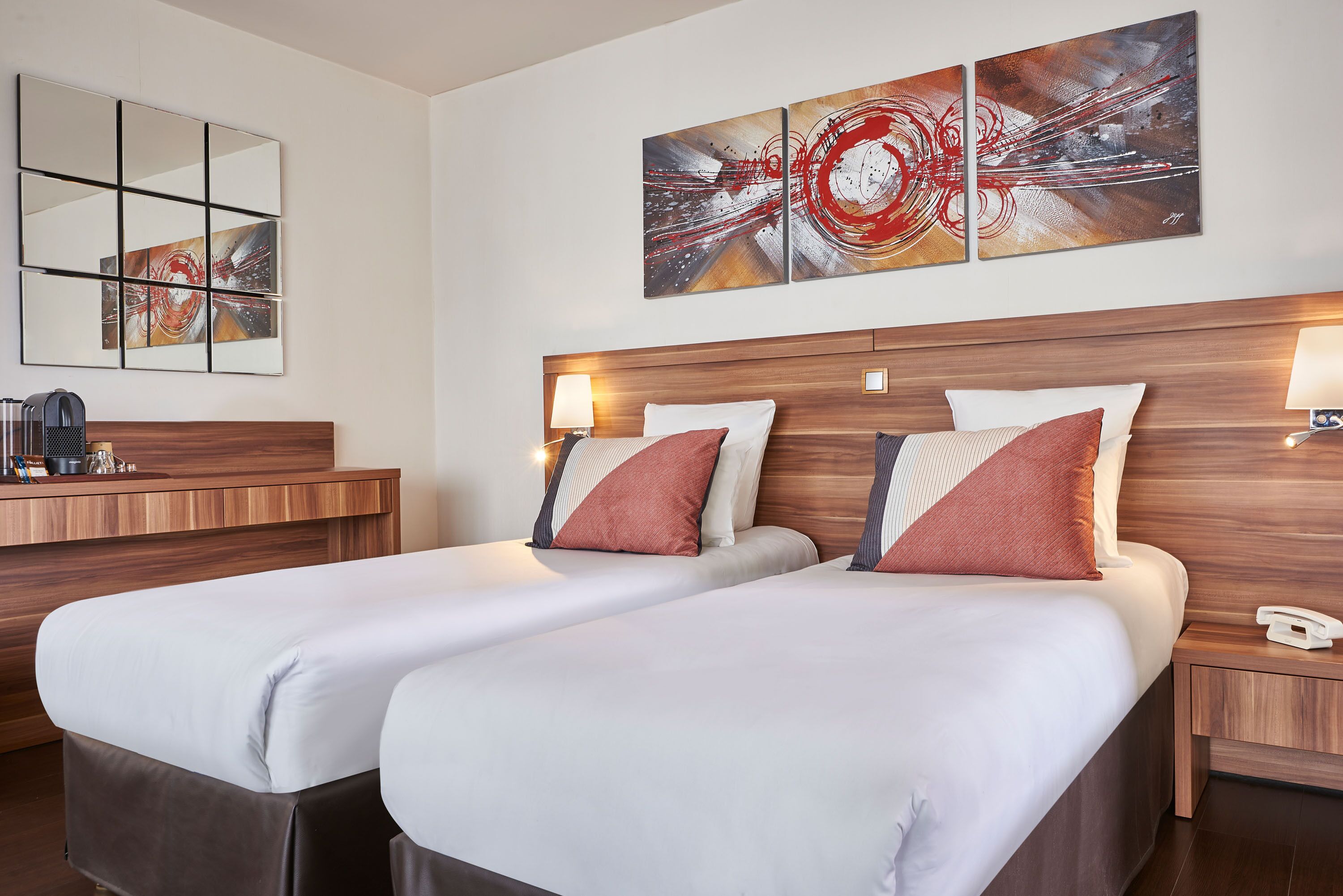 A hotel room with two beds, a wooden headboard, a bedside table with a lamp, and a large abstract painting above the beds.