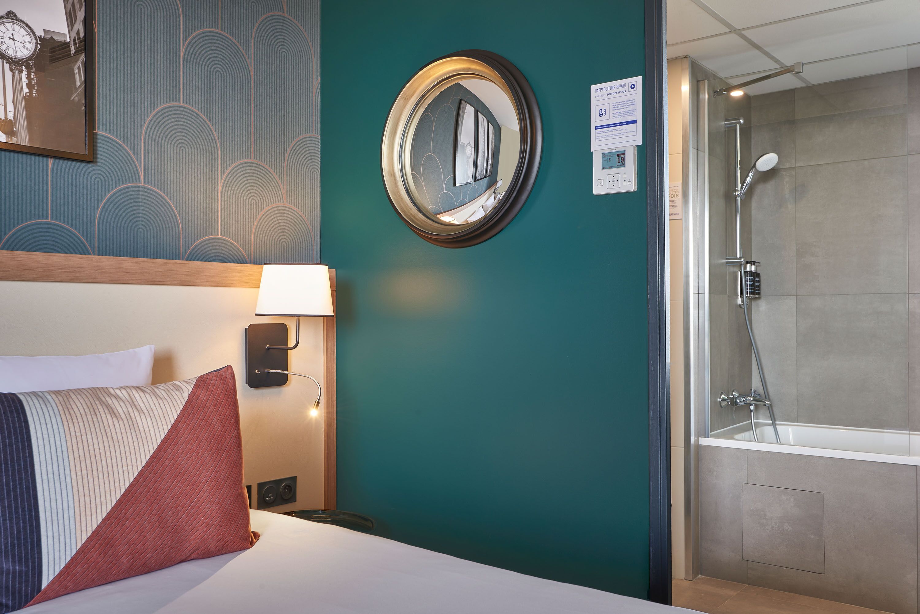 Une chambre d’hôtel comprenant un lit, une table de chevet, une horloge murale, un miroir et une salle de bain avec douche et baignoire.