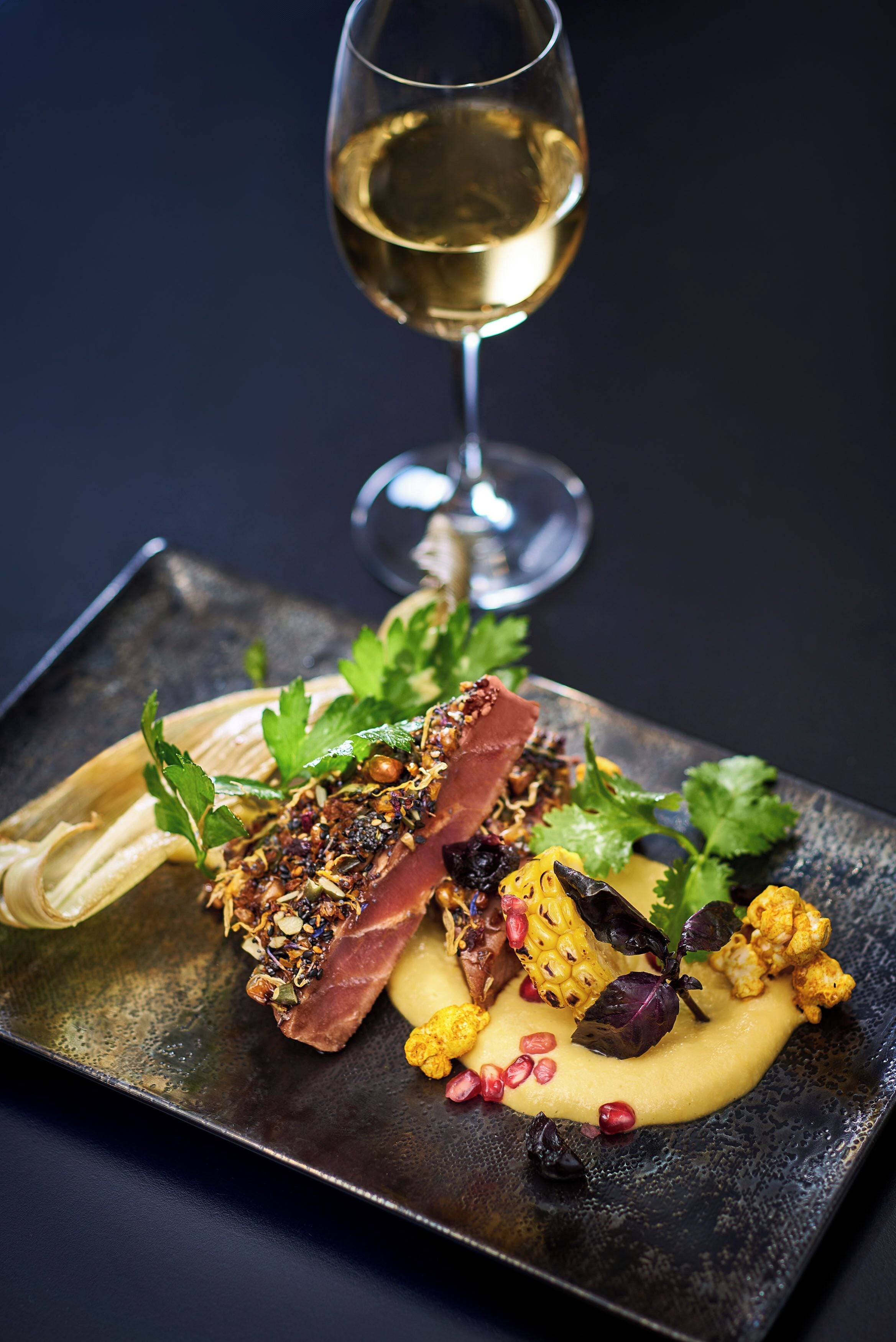 A square plate with a dish of food, a glass of white wine, and a dark background.
