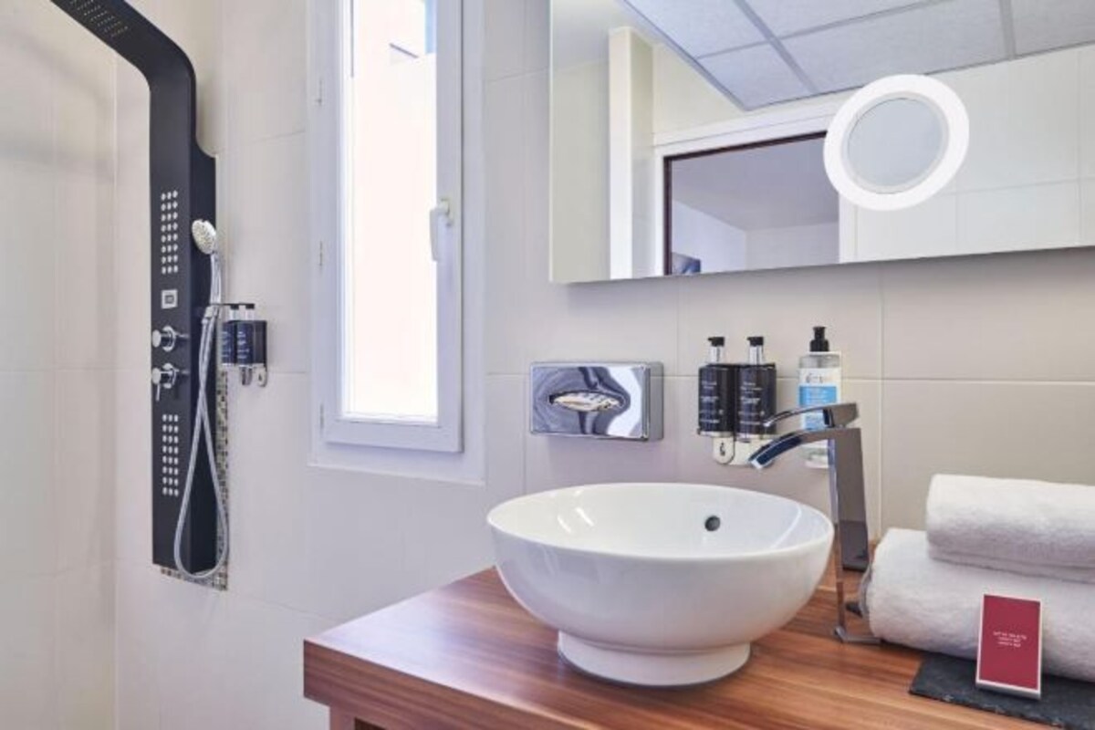 A modern bathroom with a large white sink, a handheld showerhead, a mirror with a ring light, and neatly stacked towels.
