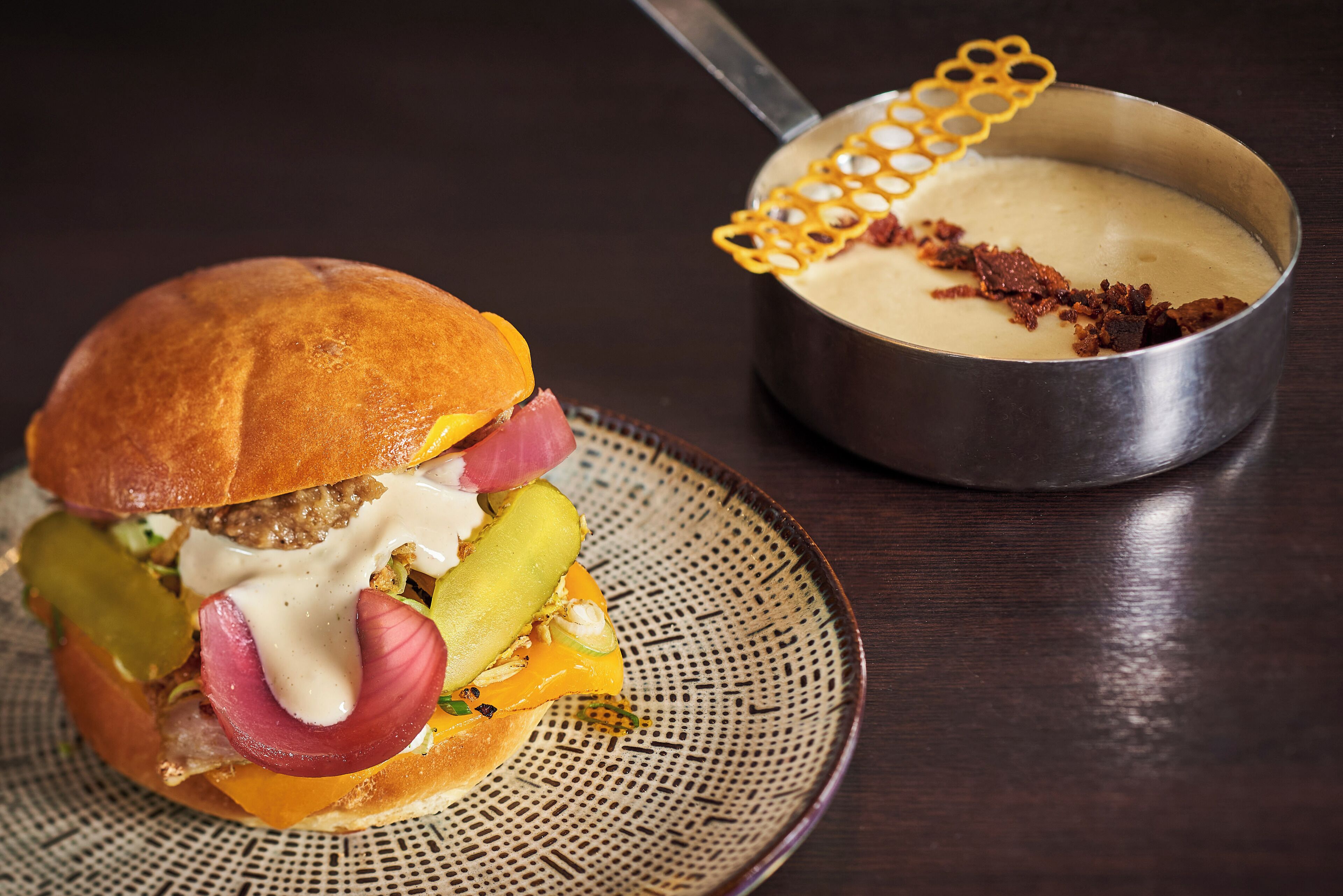 A sandwich with pickles, red onion, and cheese on a plate next to a metal pot with a honeycomb lid.