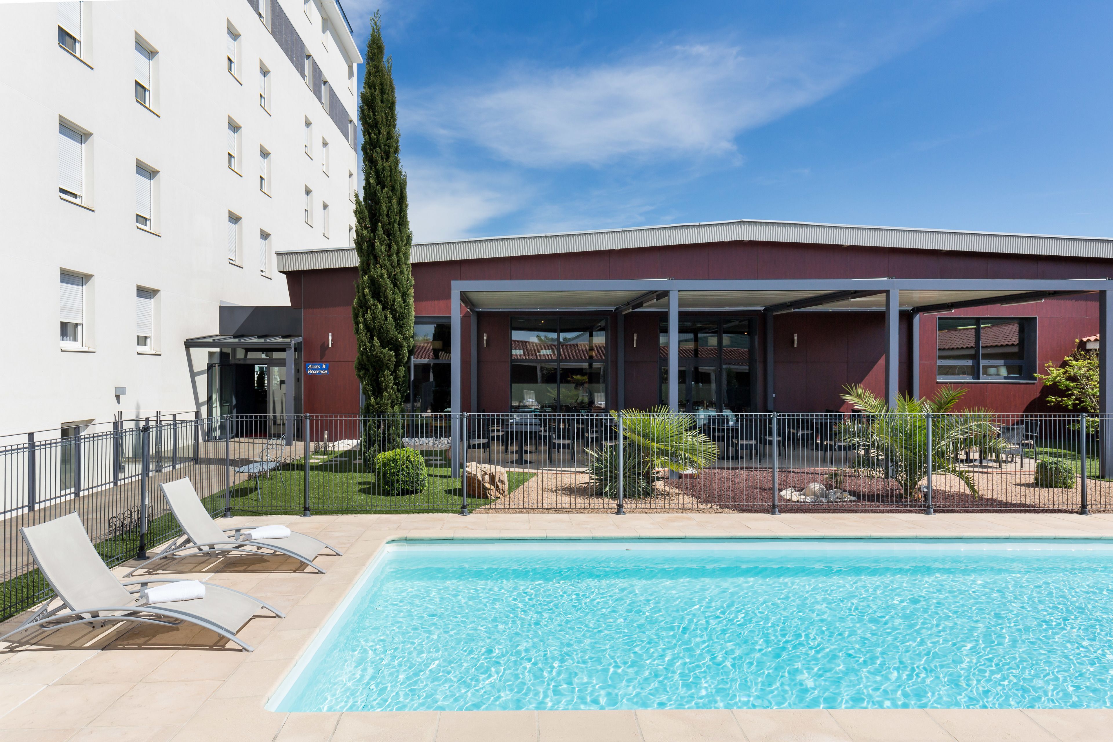 A swimming pool with lounge chairs, a covered seating area, and a building with a red facade.