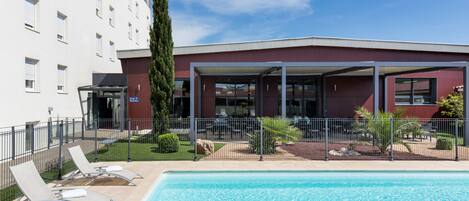 A swimming pool with lounge chairs, a covered seating area, and a building with a red facade.