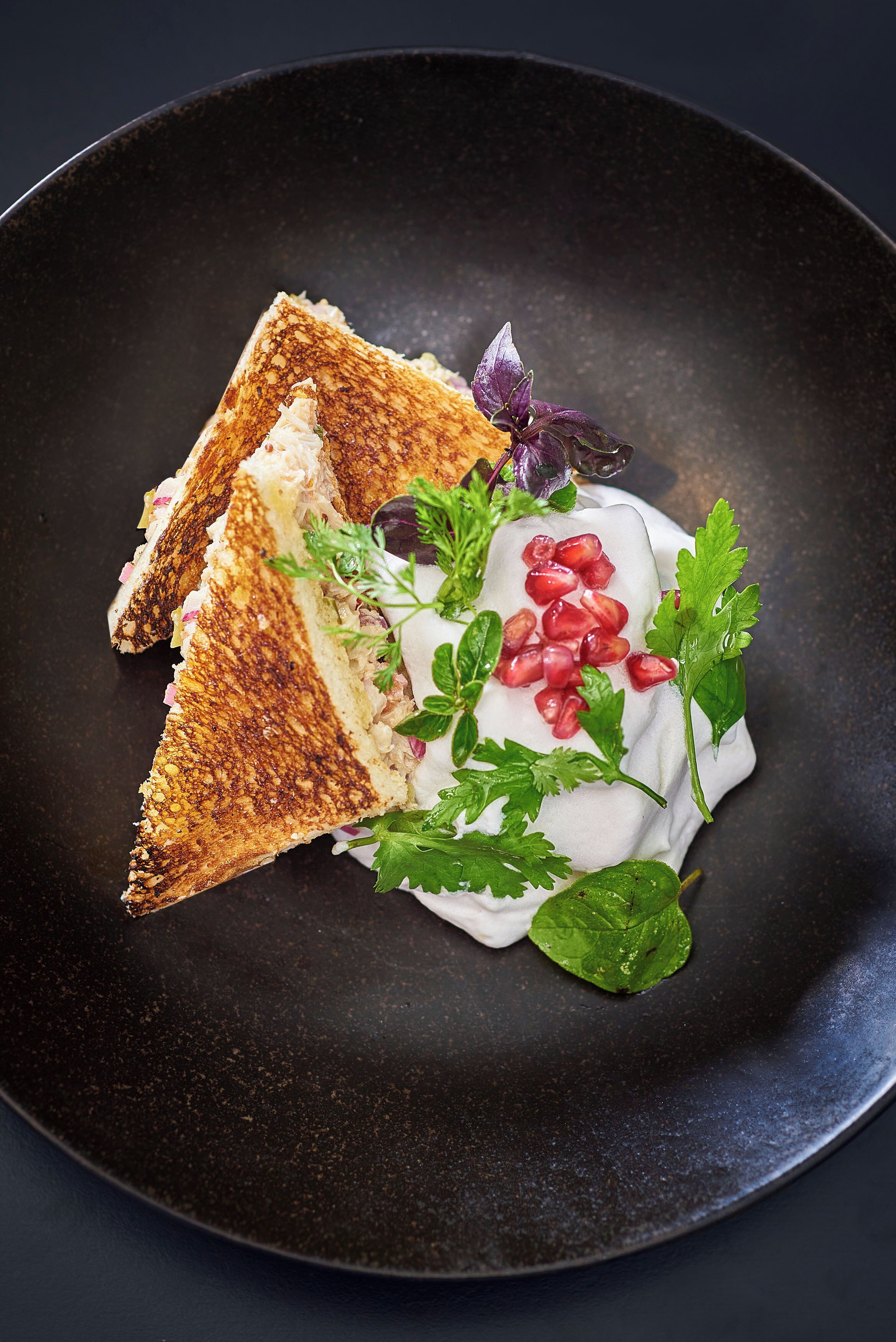 A black plate with a sandwich, pomegranate seeds, and greens.