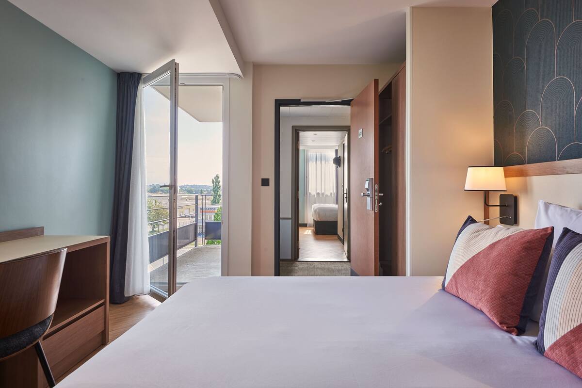 A hotel room with a bed, a desk, and a chair. There is a balcony with a view of the city.