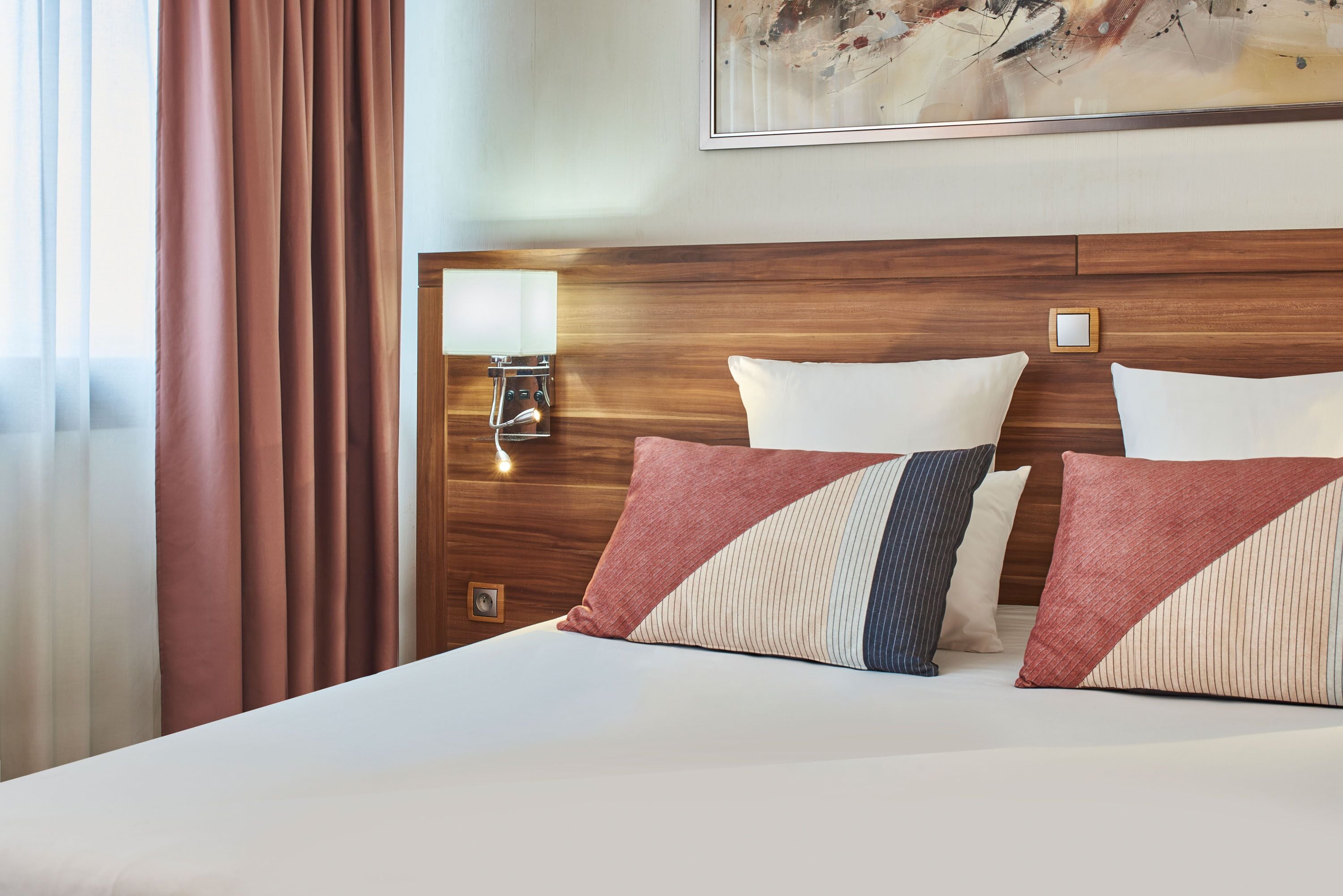 A hotel room with a wooden headboard, a bedside table with a lamp, and a large abstract painting on the wall.
