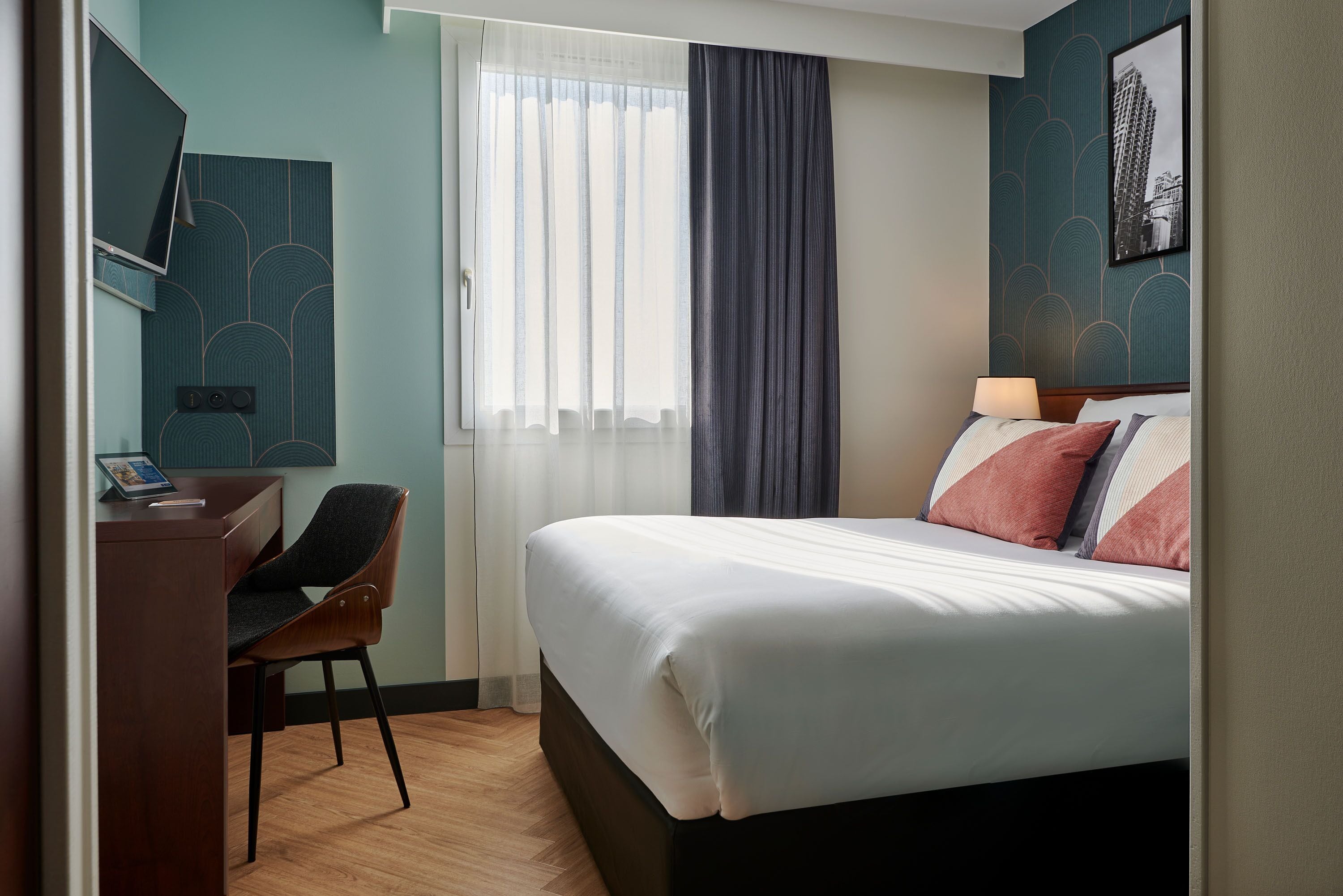 A hotel room with a bed, a desk, a chair, a window with curtains, and a framed picture on the wall.