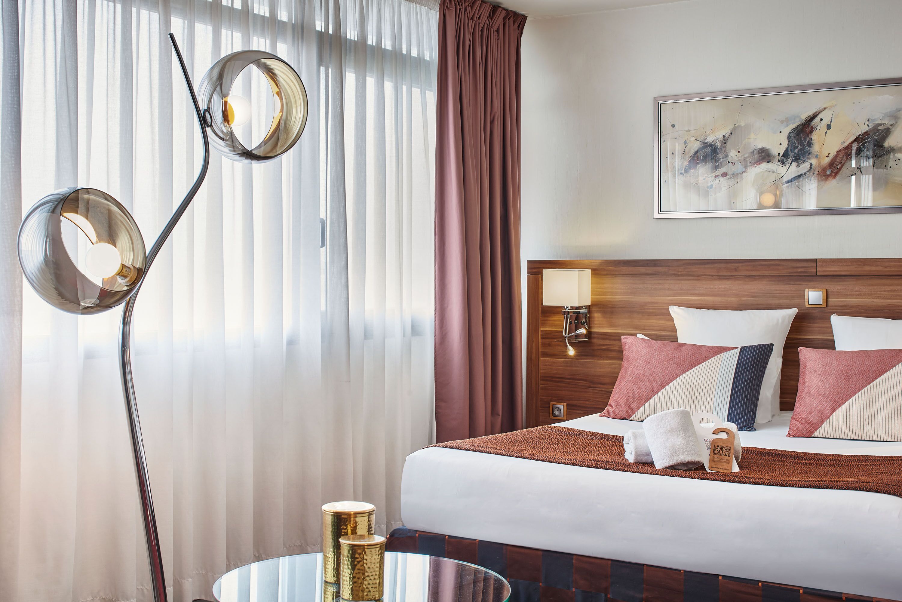 A hotel room with a bed, a bedside table with a lamp, a painting on the wall, and a window with curtains.