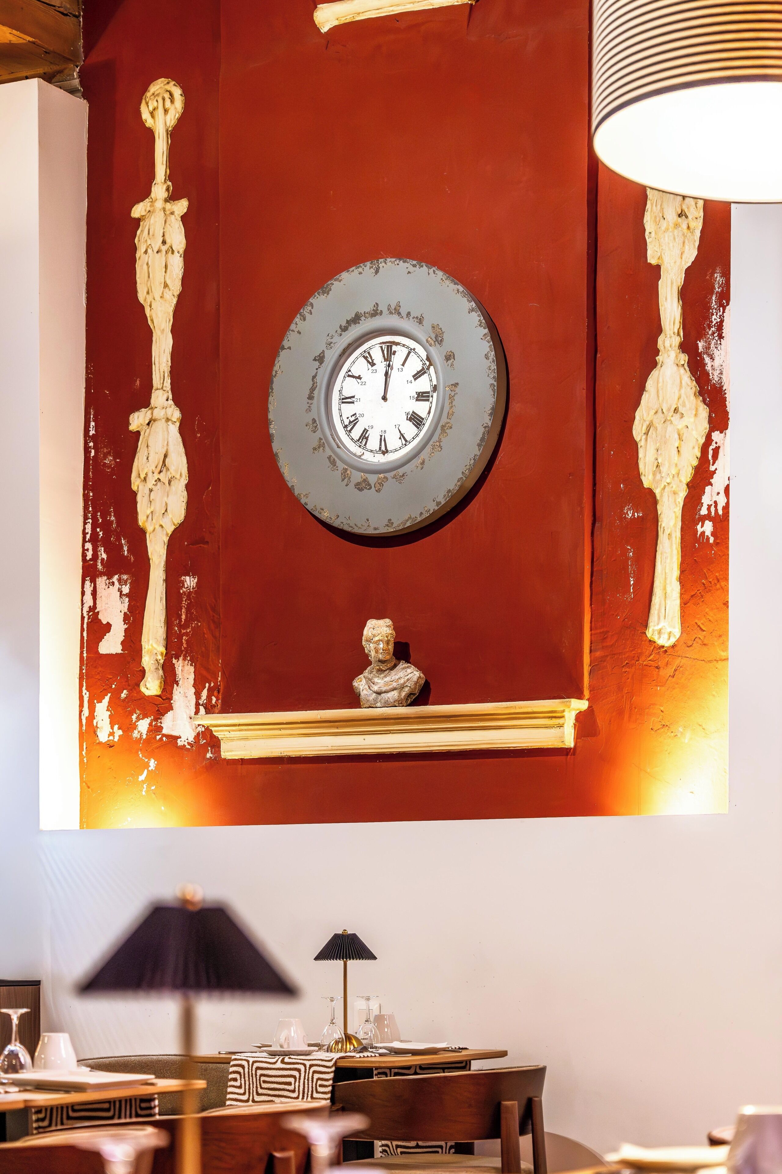 Een kamer met een rode wand, een klok, een buste en een tafel met glazen en een lamp.