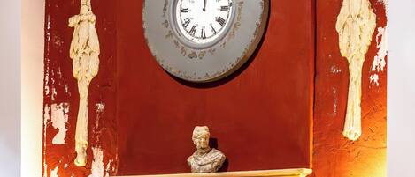 Une pièce avec un mur rouge, une horloge, un buste sculpté et une table avec des verres et une lampe.