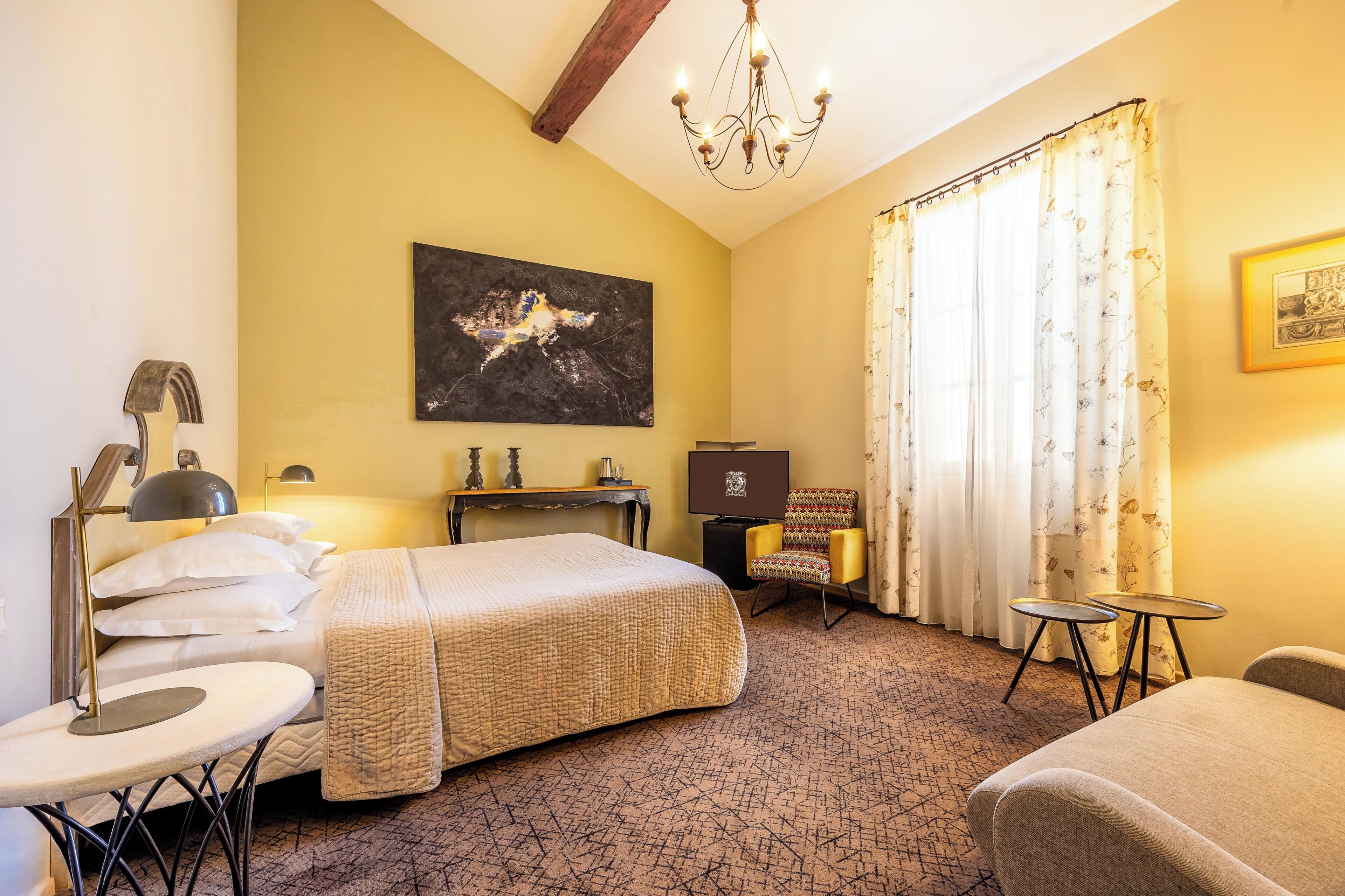 Une chambre d’hôtel comprenant un lit, un bureau, une chaise, une table, un lustre et des œuvres d’art accrochées aux murs.